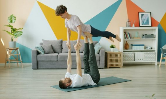 Happy couple is doing pair yoga at home, man is holding woman with legs on floor, girl is stretching arms. Relationship, active people and sports concept.